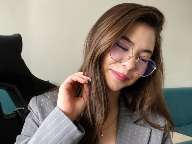 EmeraldCrivaro with brown hair, wearing a pinstripe blazer, purple-tinted glasses, and pink lipstick, touching her ear with a subtle smile, looking slightly to the side.