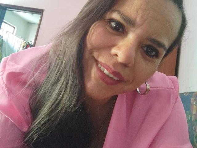VeraCruz tem cabelos castanhos longos, veste uma camisa rosa, usa brincos de argola e batom rosa, e sorri levemente inclinada, mostrando os dentes e um rosto fofo e rechonchudo..