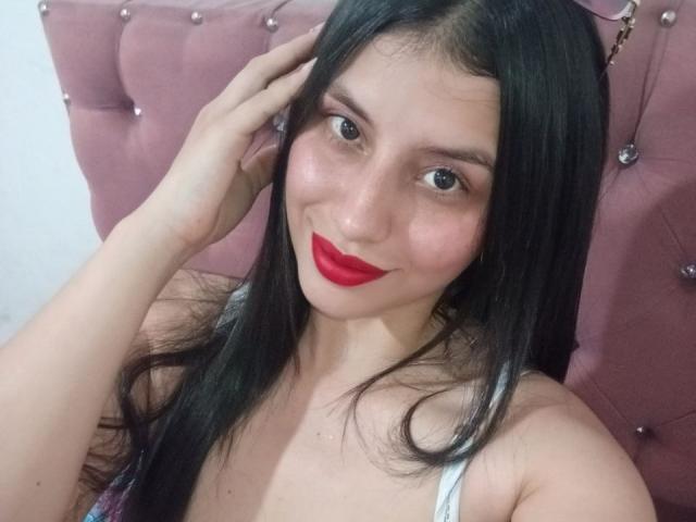 MadissonFontaine with long black hair, red lipstick, and white tank top, smiling, hand in hair, pink tufted headboard, visible tits, soft pink skin, cute face.