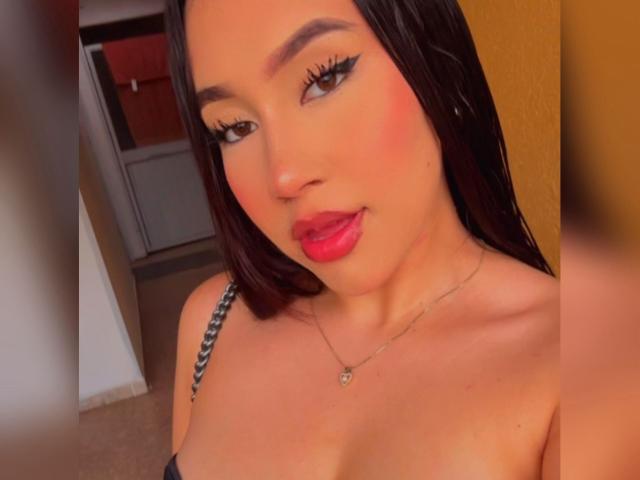 BibiSugarX has dark brown hair, red lipstick, and is showing her tits with a slight smile, wearing a necklace and black strap. Her makeup is bold with winged eyeliner.