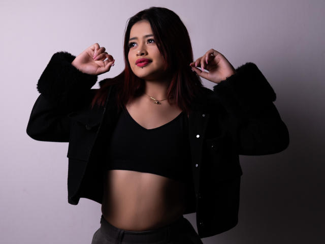 JennieRider con cabello rojo oscuro, top corto negro, chaqueta negra con mangas de piel, pantalones grises y lápiz labial rosa, manos cerca de la cabeza, sonriendo ligeramente, piel clara, linda..