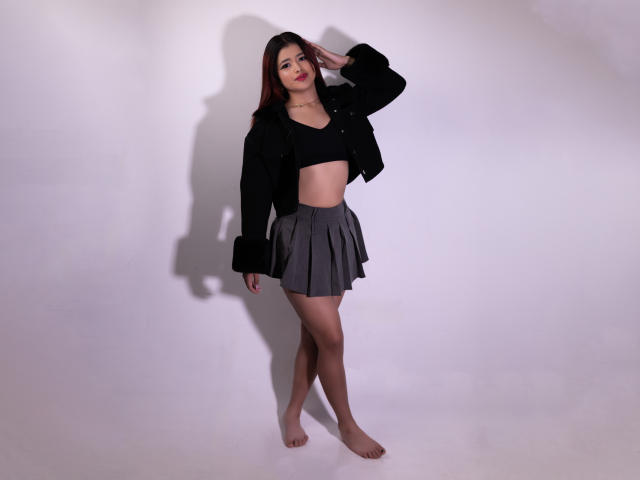 JennieRider with medium tits, wearing a black crop top, black jacket, and gray pleated skirt, standing barefoot, saluting with her right hand, shadow on white wall.