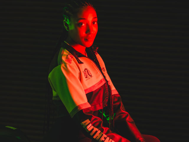 EsmeraldaRoe with red and green lighting, wearing a white and brown sports jacket, black braids, and a serious expression, sitting in a dark, shadowy environment.