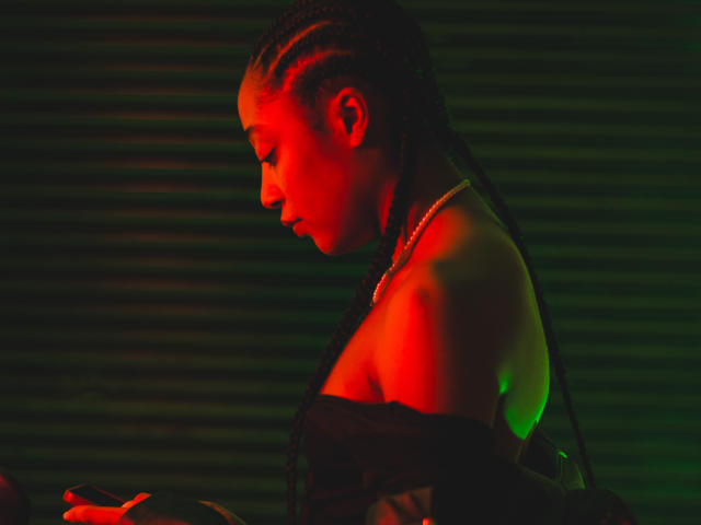 EsmeraldaRoe with braided hair, red and green lighting, holding something in her hand, off-shoulder top, necklace, side profile, looking down, sexy, dark background.