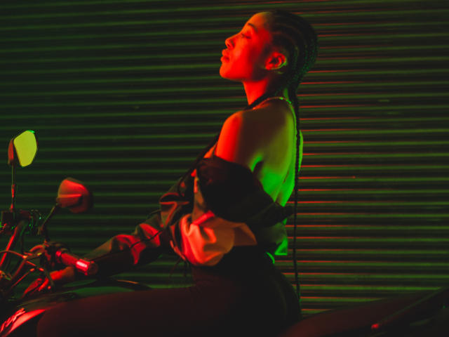 EsmeraldaRoe, red-lit, seated on a motorcycle, black off-shoulder top, green and red lighting, braided hair, side profile, holding handlebars, ribbed background, sexy, confident, stylish.