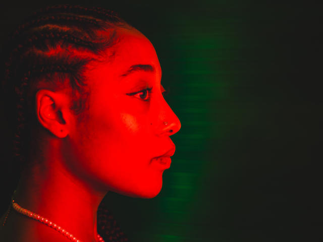 EsmeraldaRoe's red-lit face, nose ring, and braided hair in profile. She's wearing a pearl necklace, looking serious with subtle green light in the dark background.