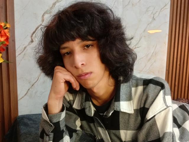 KiraFelot with curly black hair, brown eyes, and medium skin tone, wearing a black and white checkered shirt, resting their head on their hand, looking bored.
