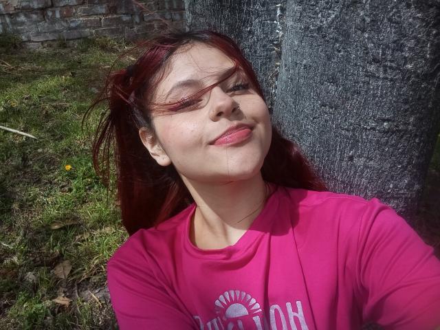 Sophie Hill, con i capelli rossi, una maglietta rosa, sorridente, con gli occhi chiusi, illuminata dal sole, in piedi di fronte a un albero, sull'erba e contro un muro di mattoni. La sua maglietta rosa mostra un disegno del sole. È carina..