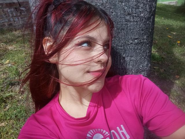 Sophie Hill mit roten Haaren, einem rosa Hemd und einem leichten Lächeln, die Augen blicken nach rechts, der Wind weht durch ihre Haare, sie steht vor einem Baum, im Hintergrund sind Gras und gelbe Blumen zu sehen..