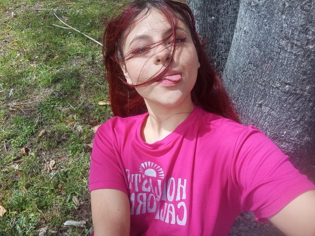 Sophie Hill, con i capelli rossi, una maglietta rosa, la lingua fuori, il labbro superiore sporgente, seno di media grandezza, un albero a destra, terreno erboso, luce solare, capelli corti, selfie casuale, leggera brezza..