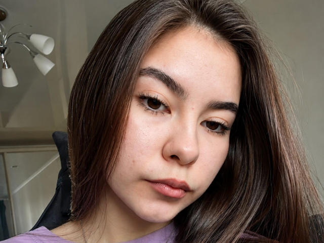 EmeraldCrivaro has brown eyes, long straight brown hair, and a neutral expression with slightly parted pinkish lips. She's wearing a purple shirt. Her skin is light with a few small blemishes.