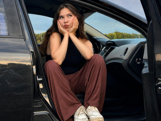 EmeraldCrivaro sitting in a car, hands on cheeks, looking to the side, in a black sleeveless top, brown pants, and white sneakers, with a serious expression.