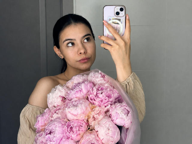 EmeraldCrivaro with dark hair, holding pink peonies, wearing off-shoulder beige top, taking mirror selfie with iPhone, looking to the side, pink nails, subtle makeup.