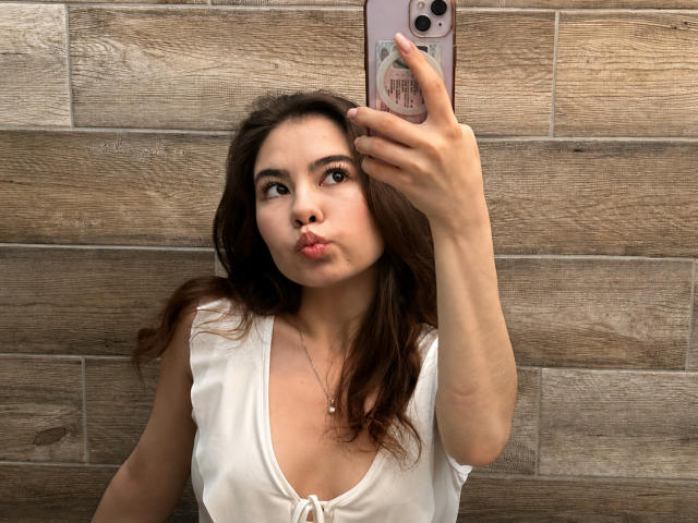 EmeraldCrivaro taking a selfie, pouting with her lips, in a white top, holding a pink phone, brown wavy hair, wooden wall background, medium tits, cute face, subtle makeup.