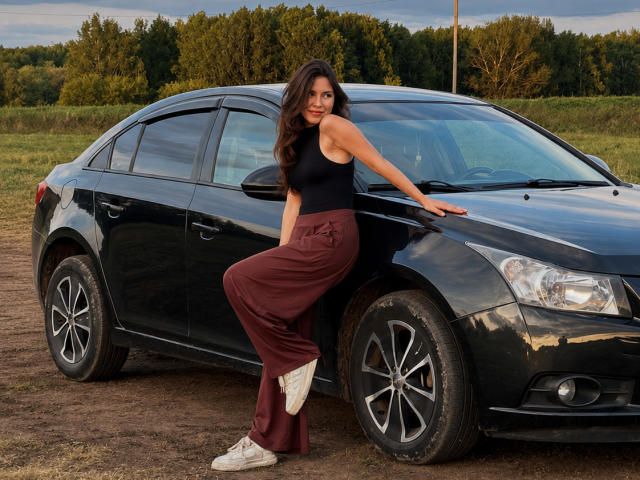 EmeraldCrivaro leans against a black sedan, wearing a black tank top and maroon pants, with white sneakers, in a field with trees in the background.