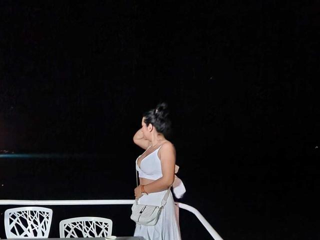EmaFoxy, avec des cheveux foncés relevés, portant un crop top blanc et une jupe, debout sur un bateau la nuit, tenant un sac, regardant sur le côté..