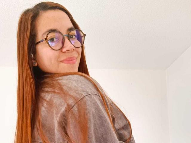 MadisonKoper with long brown hair, wearing glasses and a grey shirt, smiling slightly, showing a cute face, and looking over her shoulder.