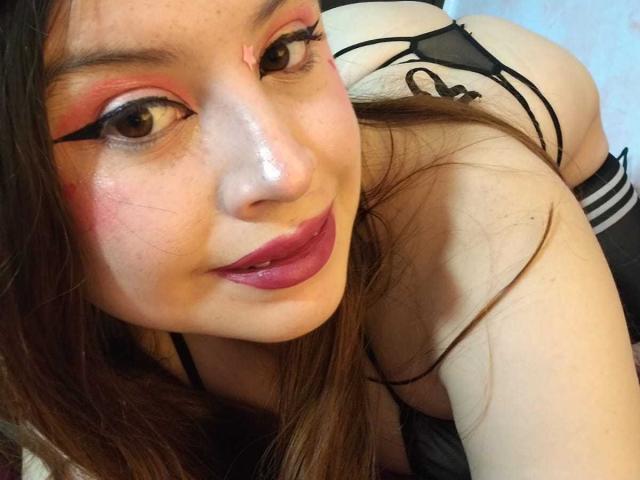 Melani Becker, con ombretto rosso, eyeliner nero e rossetto rosa, è china in avanti, mostrando il sedere in biancheria intima nera, sorridendo alla fotocamera, con lunghi capelli castani..