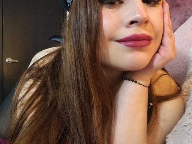 Melani Becker, con i suoi lunghi capelli castani, il rossetto rosa e il braccialetto nero, appoggia il mento sulla mano e sorride dolcemente. Il suo seno è parzialmente visibile, rivelando un accenno di scollatura..
