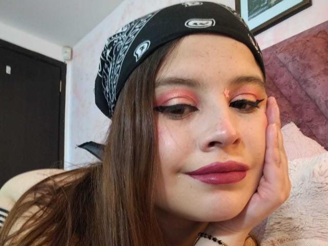 Melani Becker con ombretto rosa, bandana nera, lunghi capelli castani, labbra rosse e una mano sul viso, che mostra le sue lucide labbra e un soffice cuscino bianco..