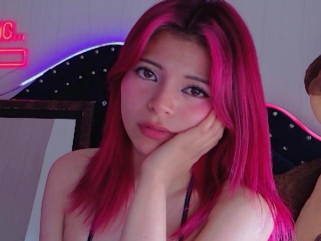 ShofiaWett con i capelli rosa, che guarda la fotocamera, con una mano sul viso, spalle scoperte, luce al neon rosa sullo sfondo, espressione delicata, pelle chiara, palpebre rosa, labbra rosa leggermente dischiuse..