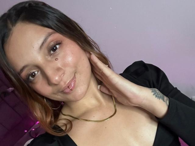SaarahJones with brown hair, nose piercing, gold necklace, black top, hand on neck, slight smile, visible tattoo on right wrist, pinkish skin, subtle makeup, medium breasts.