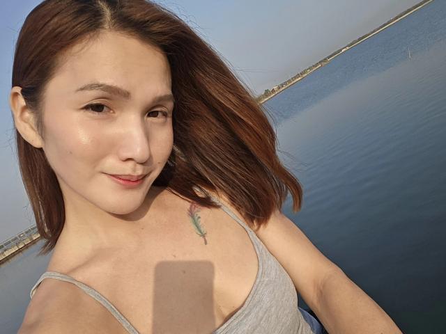 LovelyPinay69 with brown hair, light skin, small tits, and a gray tank top, smiling at the camera, showing a small tattoo on her right chest. Water background.