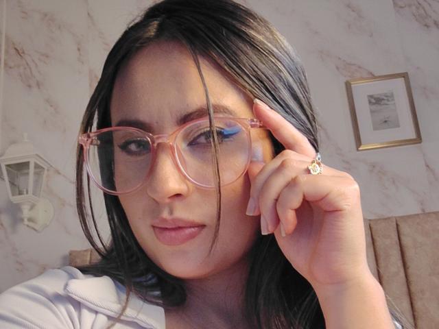 AmiiRosse with pink glasses, black hair, blue eyeshadow, white shirt, holding her cheek, wearing a ring, subtle smile, looking at camera, soft lighting, marble wall.