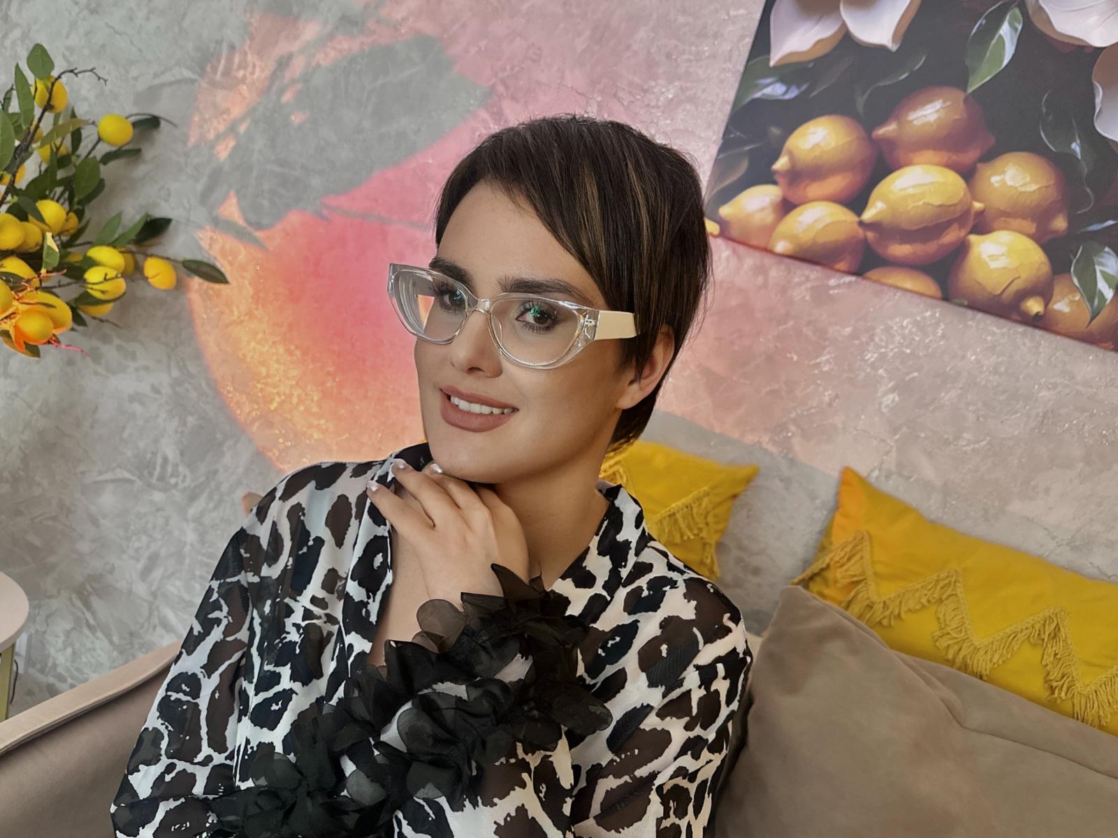 MayaNoir with short brown hair, wearing clear glasses and a black/white leopard print shirt, smiling with her hand near her chin, sitting on a yellow pillow.