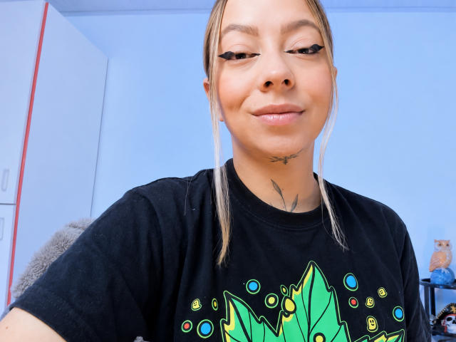 KayleeM with blonde pigtails, black t-shirt, and eyeliner, smiling at camera, small tattoo under chin, soft blue room, light skin, slight makeup, casual selfie.