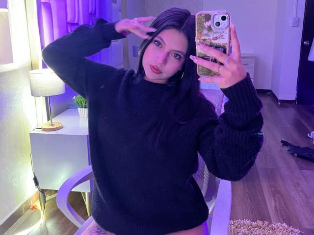 NyraVanel with long black hair, wearing a black rib-knit sweater, taking a mirror selfie with her right hand up, purple lighting, wooden floor, and a white lamp in background.