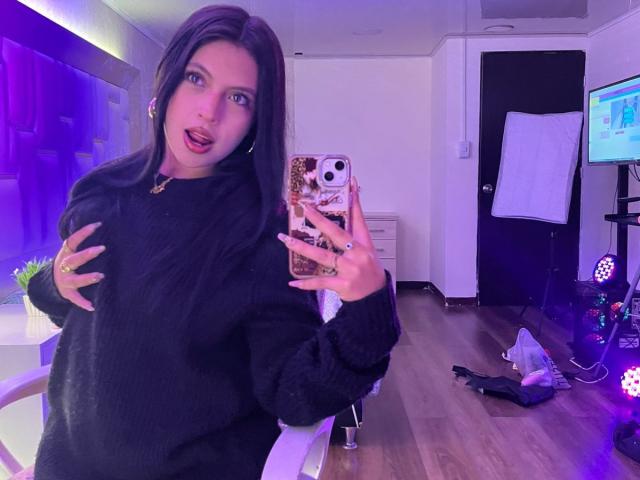 NyraVanel with long black hair, wearing a black sweater, holding a phone in front of a mirror, slightly open mouth, purple lighting, right hand on chest, wooden floor.