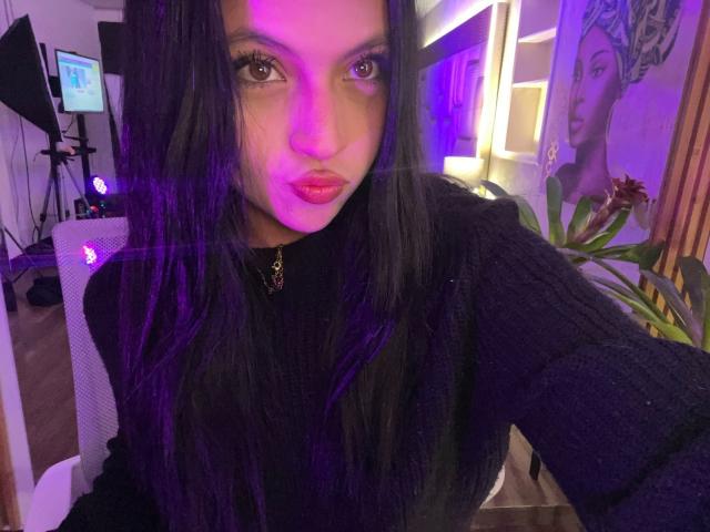 NyraVanel with long black hair, pink-purple lighting on her face, wearing a black knit sweater, pouty pink lips, and a black choker with a red gem.