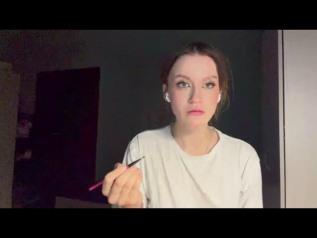 BuenaPontbriand with pale skin, brown hair in a messy bun, wearing a white shirt, holding a black eyeliner pencil, applying makeup with a focused expression.