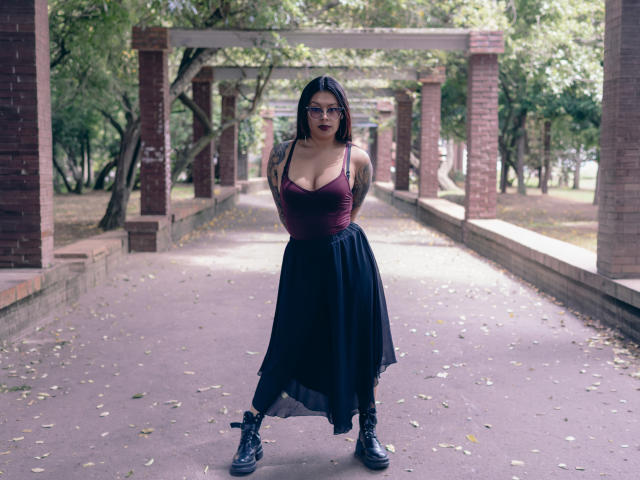 GabyJoy with big tits, wearing a maroon top, black skirt, glasses, and black boots, standing in a brick-covered path, hands behind back, looking at camera.