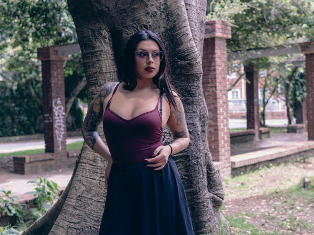 GabyJoy com batom vermelho escuro, tatuagens nos braços, vestindo uma blusa marrom e uma saia preta, em pé contra uma árvore, com as mãos na barriga, com uma expressão séria, em um parque..