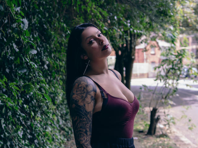 GabyJoy, con i capelli scuri, un profondo scollo in una maglia color borgogna, un braccio tatuato e rossetto scuro, in piedi accanto a una vegetazione verde. Seno, tette, che guarda in alto, strada sullo sfondo..