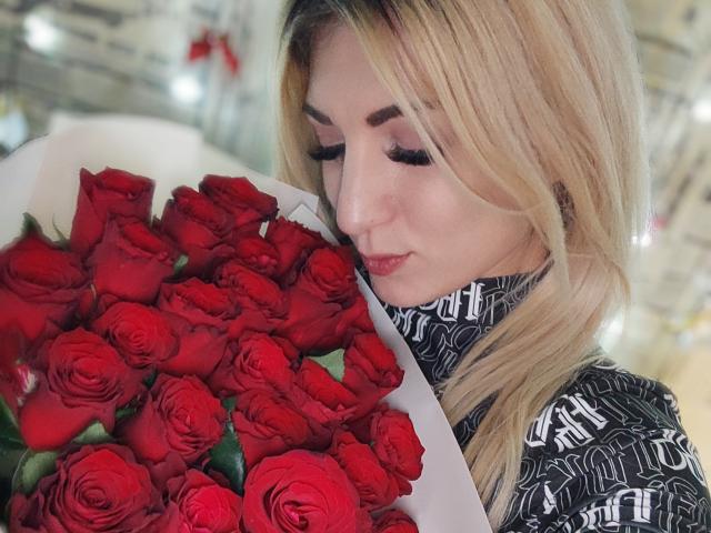 KamMlily with blonde hair, holding a bouquet of red roses, wearing a black and white patterned shirt, eyes closed, nose slightly upturned, looking cute and relaxed.