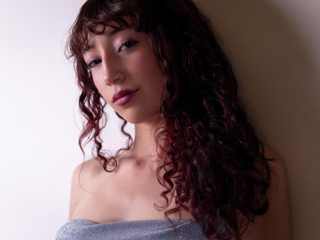 LilyDickenson with curly dark red hair, pink eyeshadow, nose ring, and purple lipstick, wearing a sparkly silver top, has a cute, sexy, and attractive face.