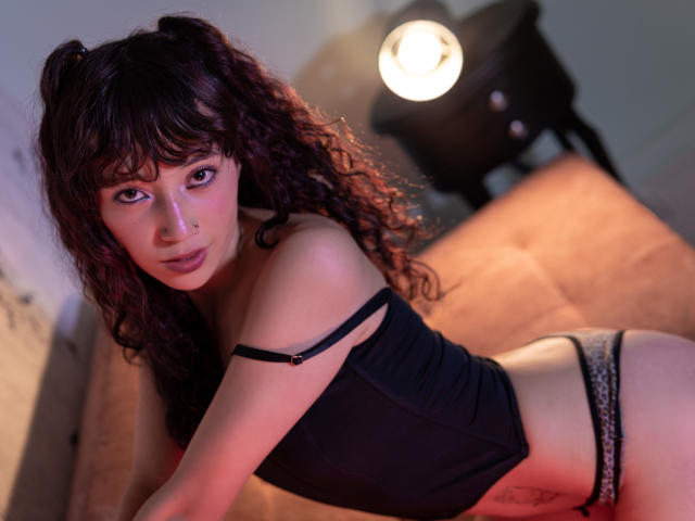 LilyDickenson with curly dark hair in pigtails, wearing a black top and leopard print panties, leaning forward on a beige sofa, looking at the camera with a seductive expression.