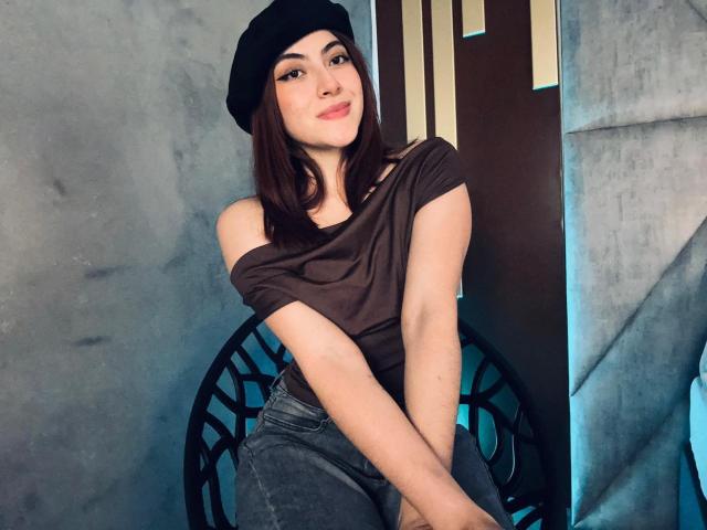 SpencerLe, com cabelo castanho escuro, gorro preto, blusa marrom com um ombro descoberto e calça jeans azul, sorrindo, com os braços cruzados, em frente a uma cadeira circular de metal preta..