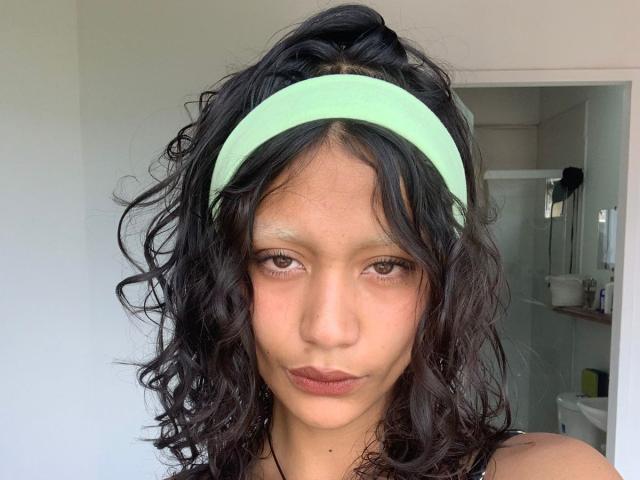 AliceFirek with curly black hair, green headband, and pouty lips, showing a neutral face. Her small brown eyes and light skin are prominent. Her ass is not visible.