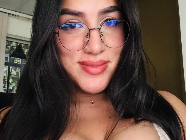 Alana Harris ha lunghi capelli neri, occhiali blu riflettenti, un anello al naso e un piercing al labbro. Indossa una maglietta bianca con scollatura pronunciata e una piccola collana..