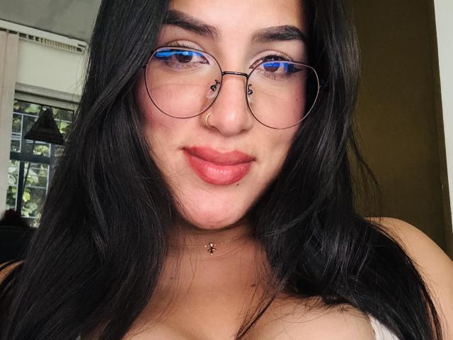 Alana Harris, con lunghi capelli neri, occhiali blu riflettenti, un anello al naso, rossetto rosa e una collana, indossa una maglietta bianca che mette in mostra il suo seno, e sorride leggermente..