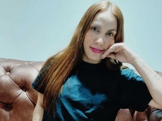 SusanThomass with long brown hair, pink lipstick, black shirt, and tattoo on right arm, resting chin on hand, sitting on brown tufted couch.