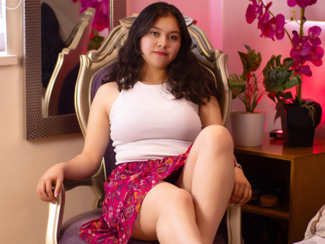 Cristine Bernard está sentada en una silla, vestida con una camiseta blanca sin mangas y una falda rosa con estampado floral, con las rodillas separadas, mostrando sus muslos. Tiene un busto de tamaño medio y cabello negro ondulado..