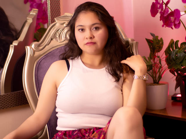 CristineBernard, a chubby Latina woman with medium tits, wearing a white tank top and pink floral skirt, sits on a purple chair, touching her hair. Pink orchids in background.