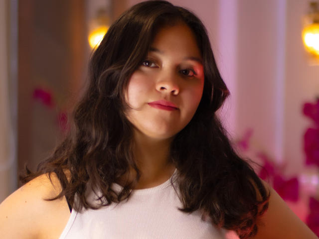Cristine Bernard a les cheveux noirs et ondulés, porte un haut blanc sans manches et du rouge à lèvres rose, et regarde légèrement sur le côté avec un sourire discret, dans une pièce éclairée chaleureusement..