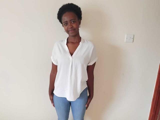SweetHeidi has dark skin, short curly black hair, small tits, wearing a white V-neck shirt and blue jeans, standing with hands in pockets, smiling slightly.