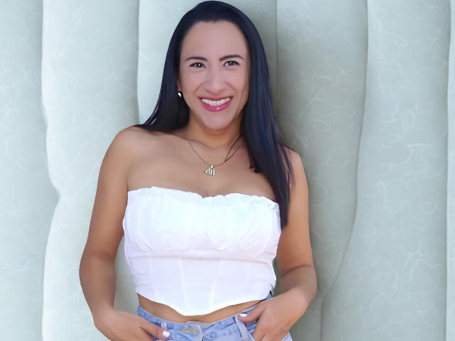 JadeFoster with black hair, smiling, wearing a white strapless top, showing small tits, and denim shorts, hands on waist, wearing a necklace, standing against a green patterned background.